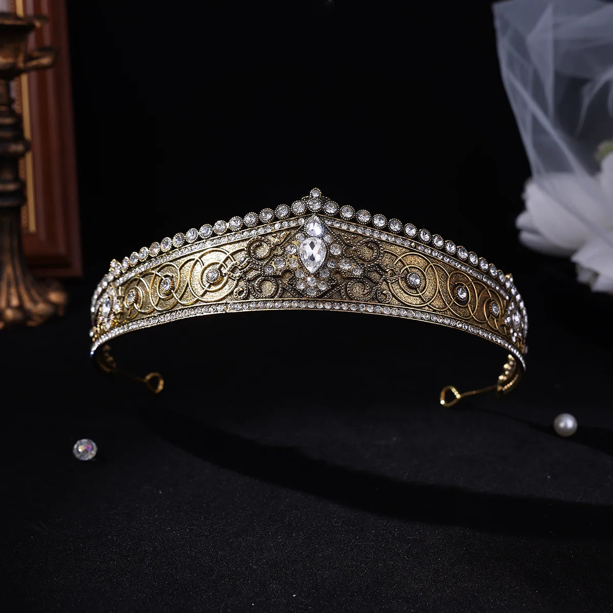 An ornate gold and silver filigree tiara crown decorated with clear rhinestones and pearl-like accents, displayed on a black background with a small loose gem and veil in the background.