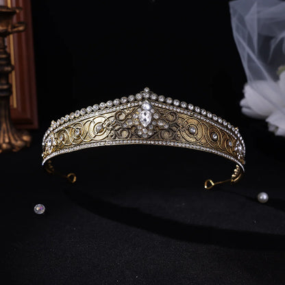 An ornate gold and silver filigree tiara crown decorated with clear rhinestones and pearl-like accents, displayed on a black background with a small loose gem and veil in the background.