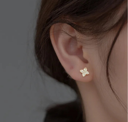 A close-up of a woman's ear wearing a small gold butterfly stud earring, with her hair, neck, and hand visible against a plain gray background.