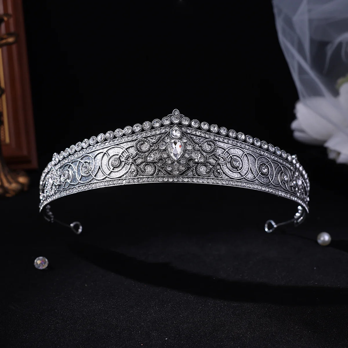 A silver-toned ornate tiara headband decorated with intricate filigree patterns and clear rhinestones, displayed on a black background with a pearl and a loose rhinestone nearby.