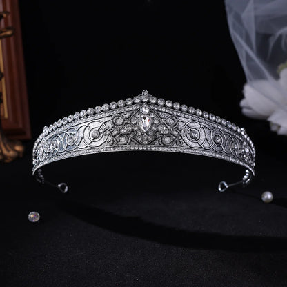 A silver-toned ornate tiara headband decorated with intricate filigree patterns and clear rhinestones, displayed on a black background with a pearl and a loose rhinestone nearby.
