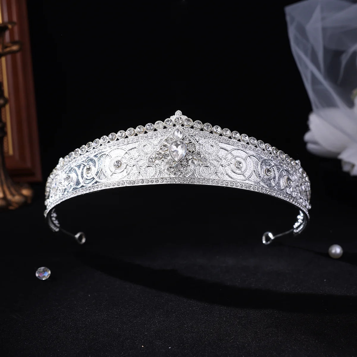 A silver tiara headband adorned with intricate floral engravings and sparkling clear rhinestones, displayed on a black background with a soft bridal veil in the background.