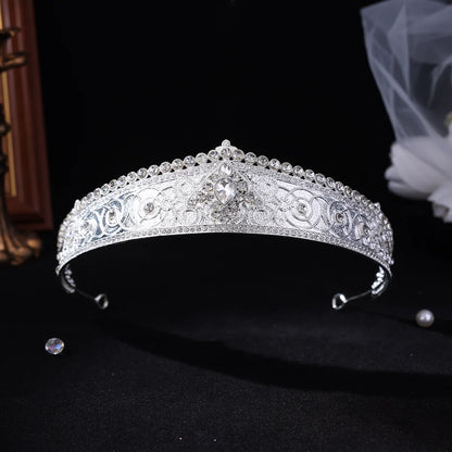 A silver tiara headband adorned with intricate floral engravings and sparkling clear rhinestones, displayed on a black background with a soft bridal veil in the background.