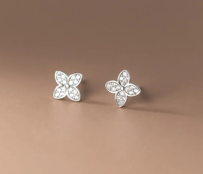 A pair of silver four-petal flower stud earrings set with clear stones, displayed on a smooth, warm brown background.