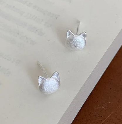A pair of silver stud earrings shaped like small cat heads with textured, matte-finish domes and pointed ears, resting on an open book with a brown wooden surface visible at the corner.