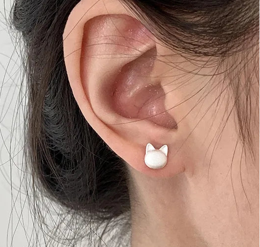 A close-up of a person's ear wearing a small white stud earring shaped like a cat head, with dark hair pulled back against a plain light background.