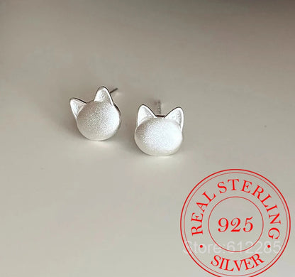 A pair of small sterling silver stud earrings shaped like cat heads, shown on a light neutral surface with a red 'REAL STERLING 925 SILVER' stamp overlay in the bottom right corner.